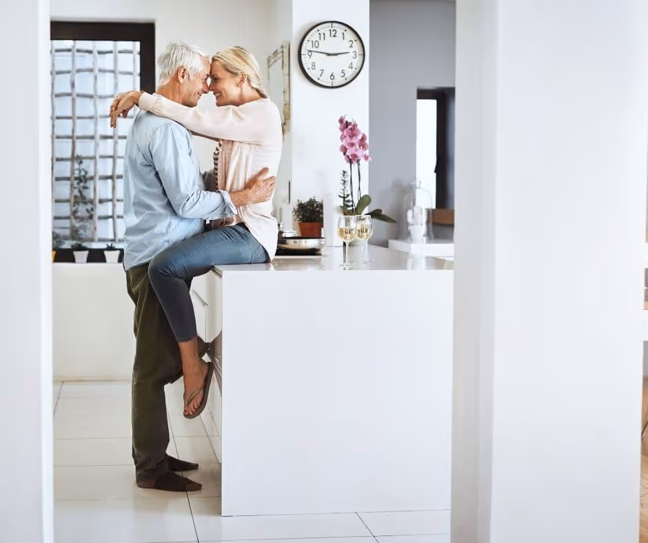 An older woman sits on the kitchen counter while an older man embraces her waist. She has her arms over his shoulders and they're heads are pressed together.