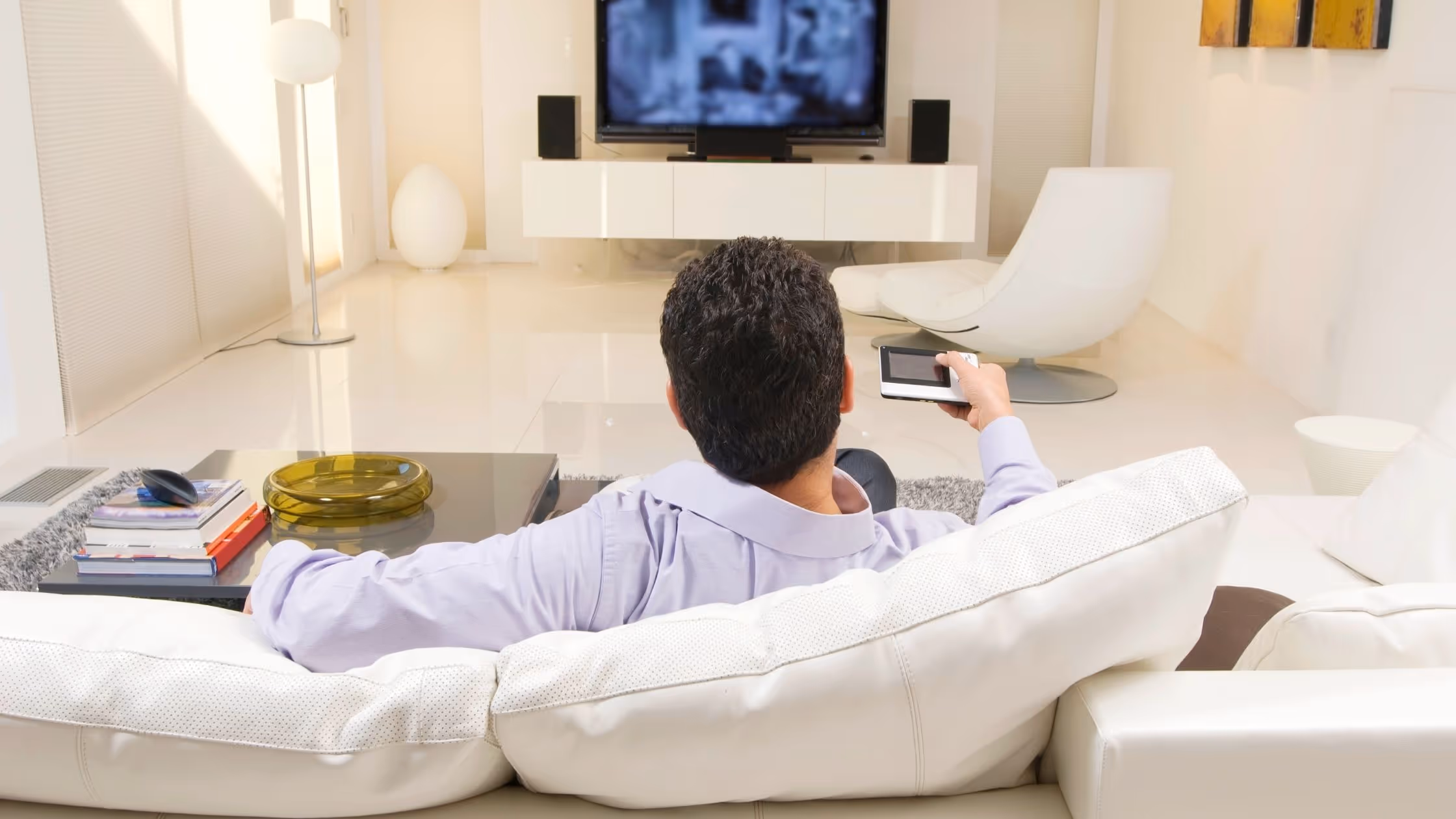 A man sits on a white leather couch, his back faces the camera, he is holding a tv remote and it is pointed at the television