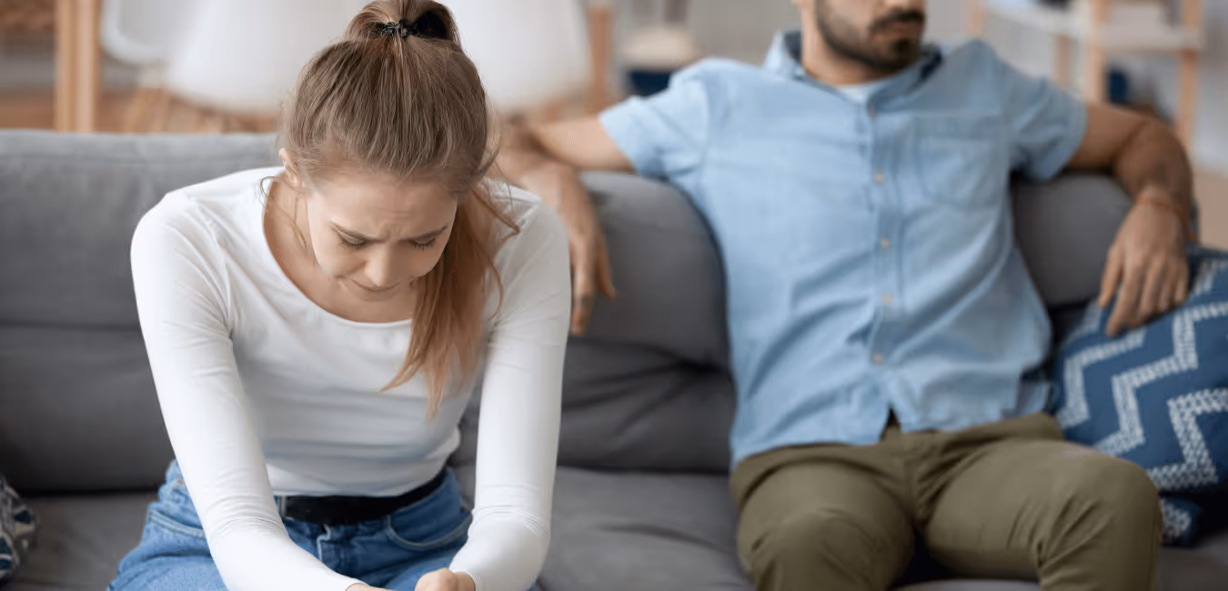 a white heterosexual couple sit on a couch. The woman is distressed and looking down, the male is leaning back on the couch looking away
