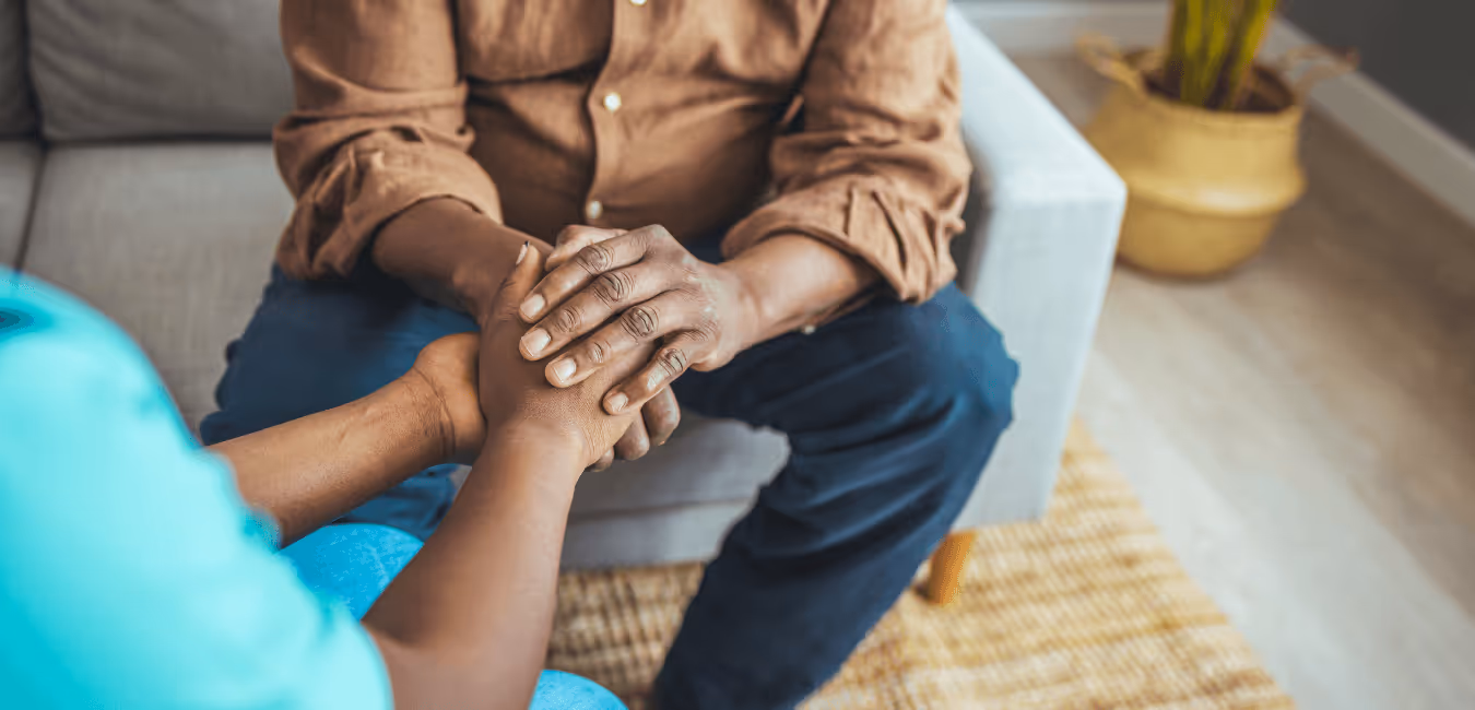 Close-up of two people seated in a room, holding hands as a gesture of comfort and support.