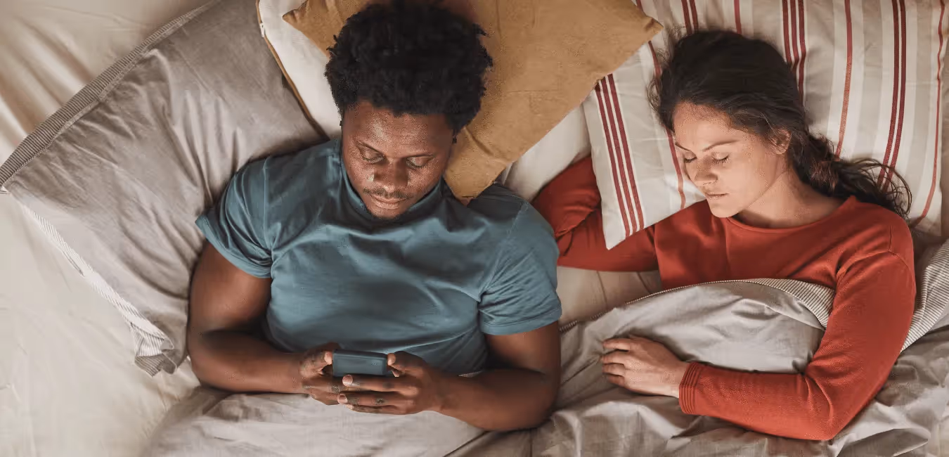 Interracial couple lying on bed and man using his phone while woman is sleeping