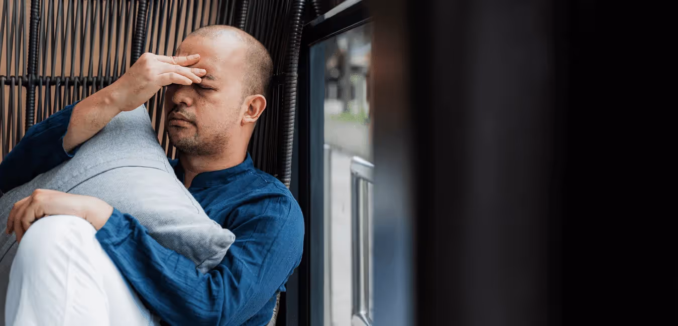 Bald man sitting near window holding pillow looking depressed at home