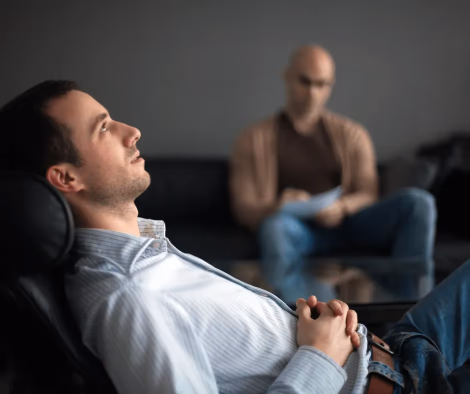 A man sits in the foreground of the photo leaning back in a chair with his hands on his stomach, another man sits on the background with a piece of paper and pen