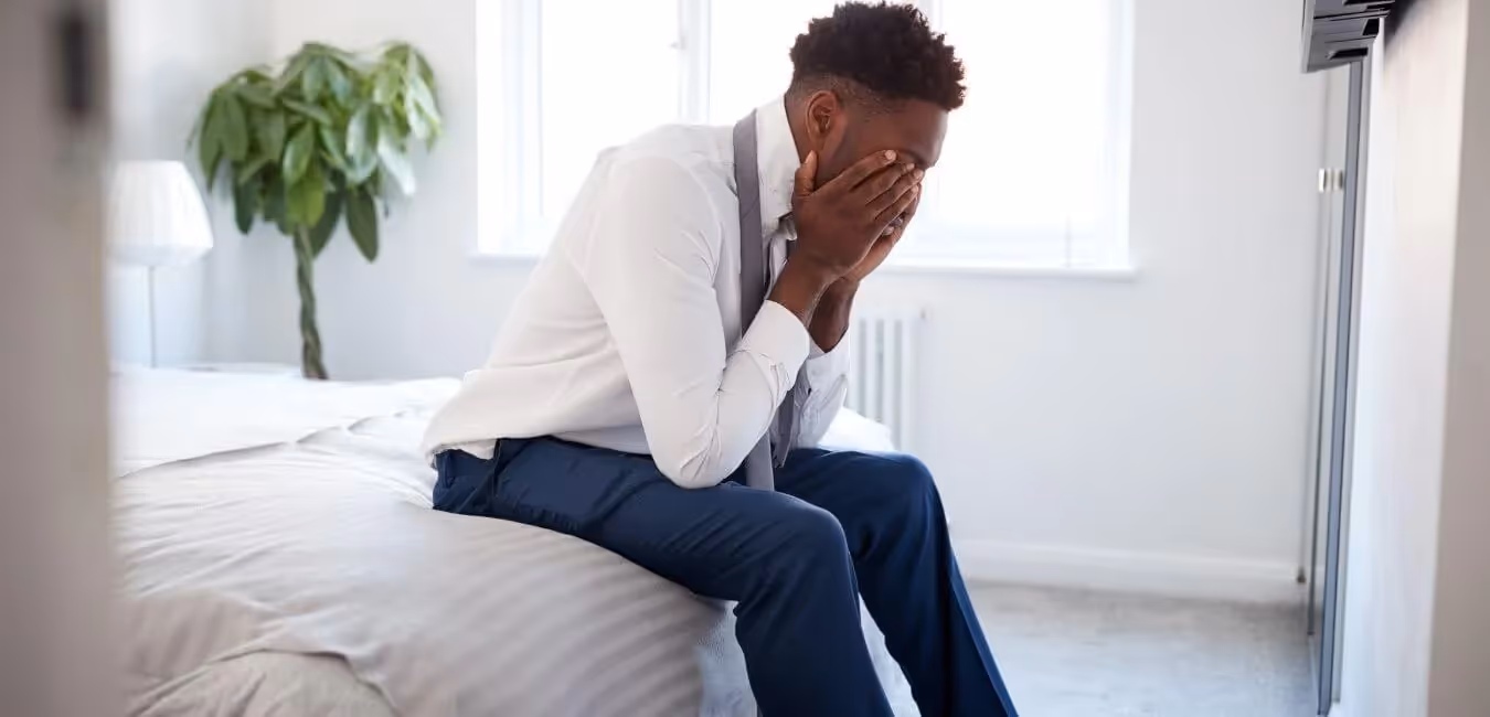 an African American man sits on the edge of a bed with his head in his hands