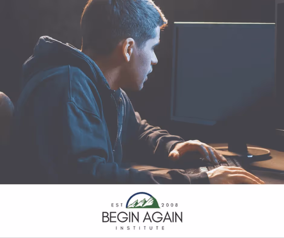 A person who identifies as male powers up a computer. The room is dark and he is wearing a hoodie.