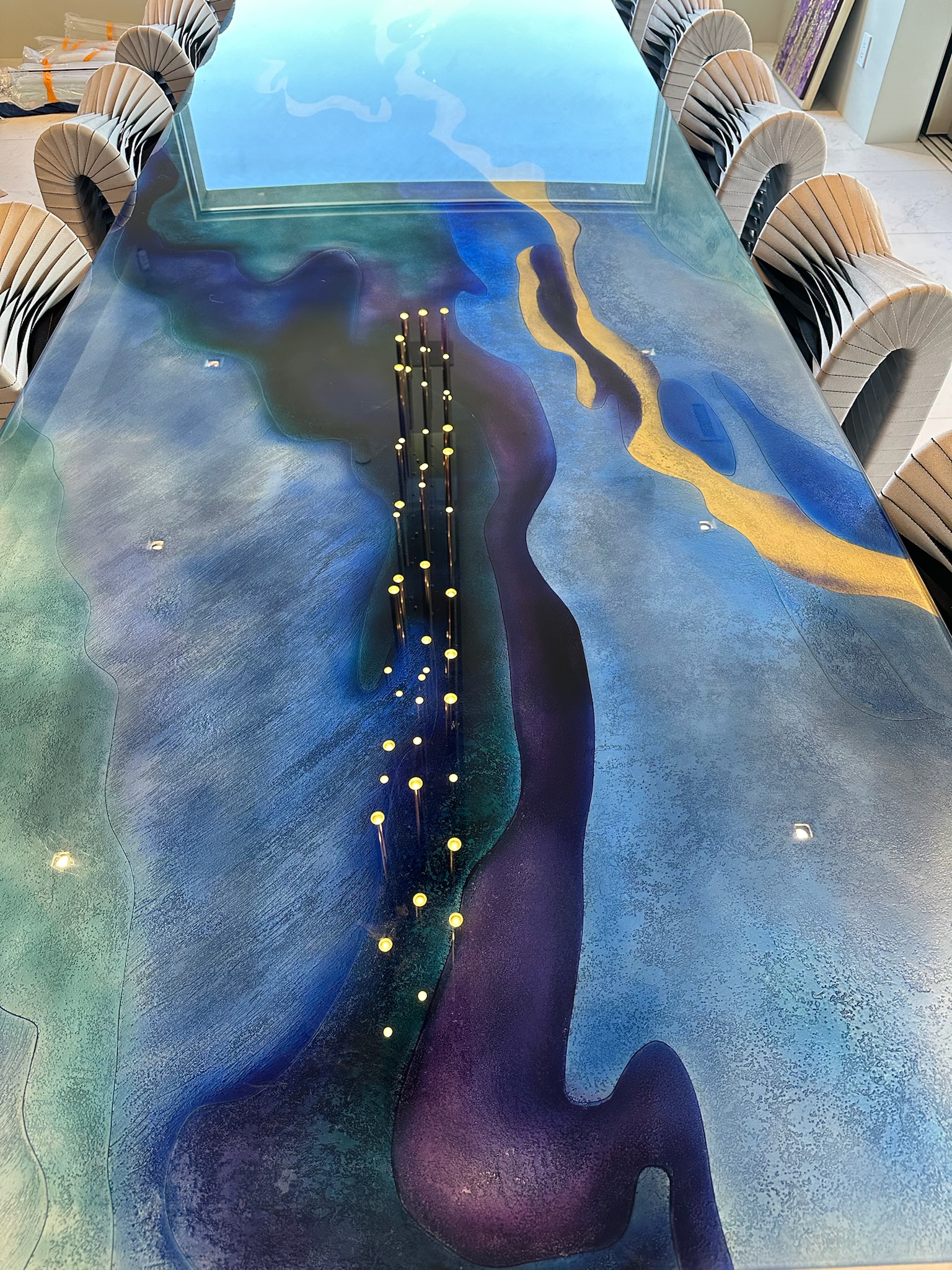 Long rectangular dining table with a glossy, hand painted glass. Artistic surface featuring abstract blue, purple, and yellow patterns resembling a river or landscape, surrounded by seven upholstered chairs on each side, with light reflections and a window reflection visible on the table surface.