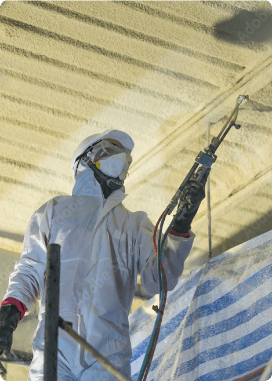 Worker in protective suit, gloves, mask, and goggles spraying insulation foam onto a ceiling.