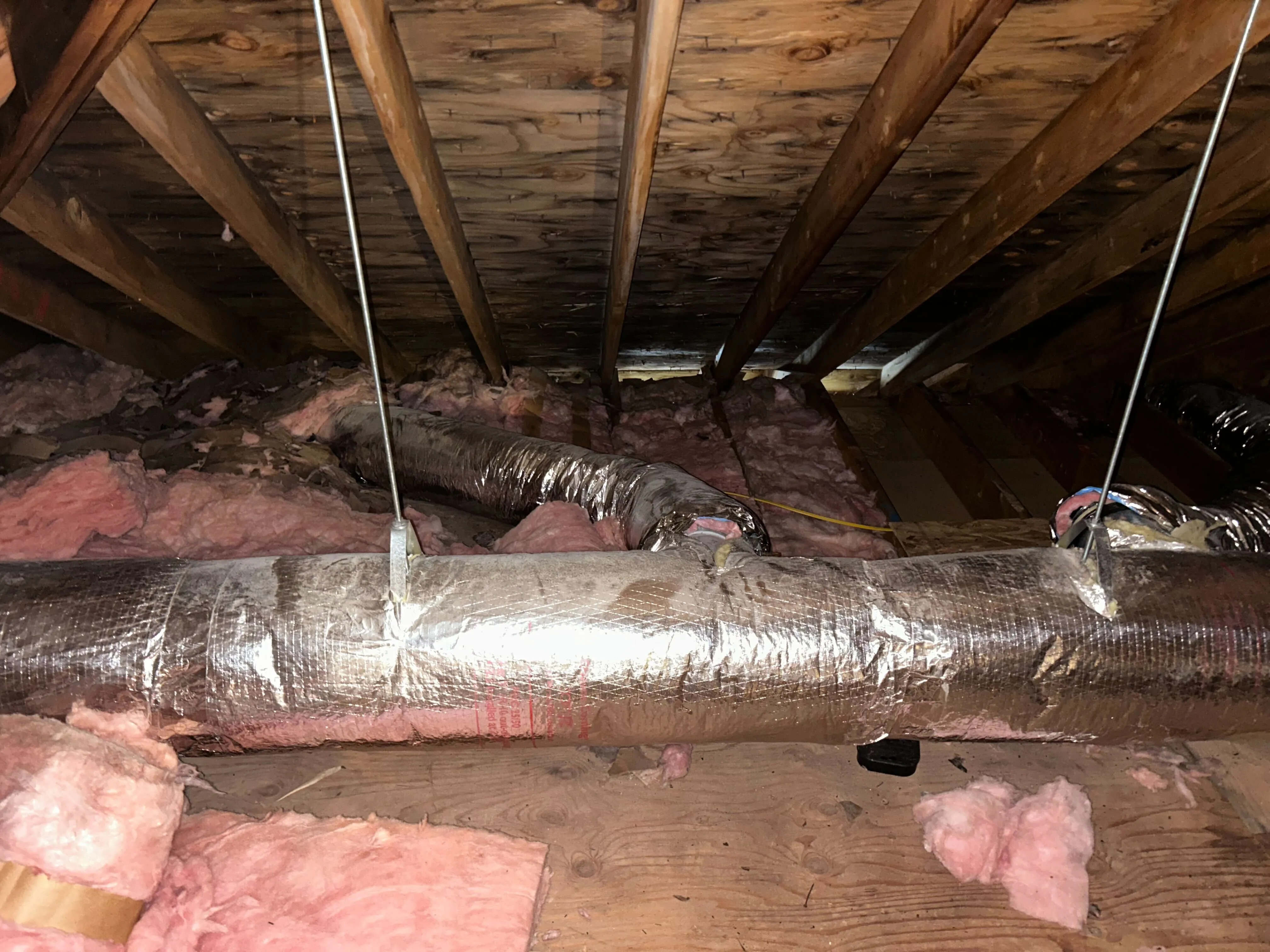 Attic with exposed wooden beams, pink fiberglass insulation, and insulated air ducts suspended from the ceiling.