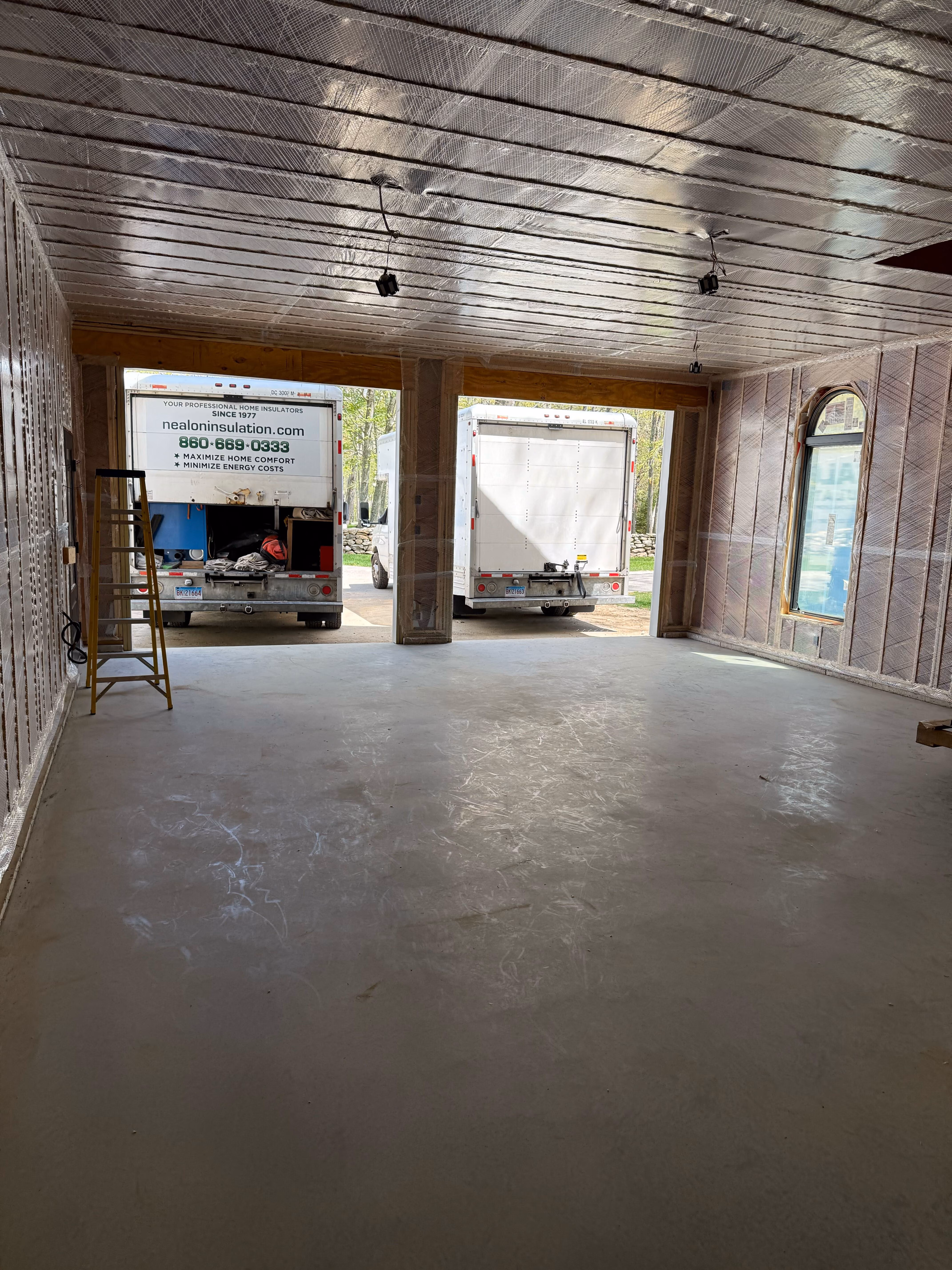 Interior of a construction site with reflective insulation installed on the walls and ceiling, two white trucks parked outside, and a yellow ladder inside.