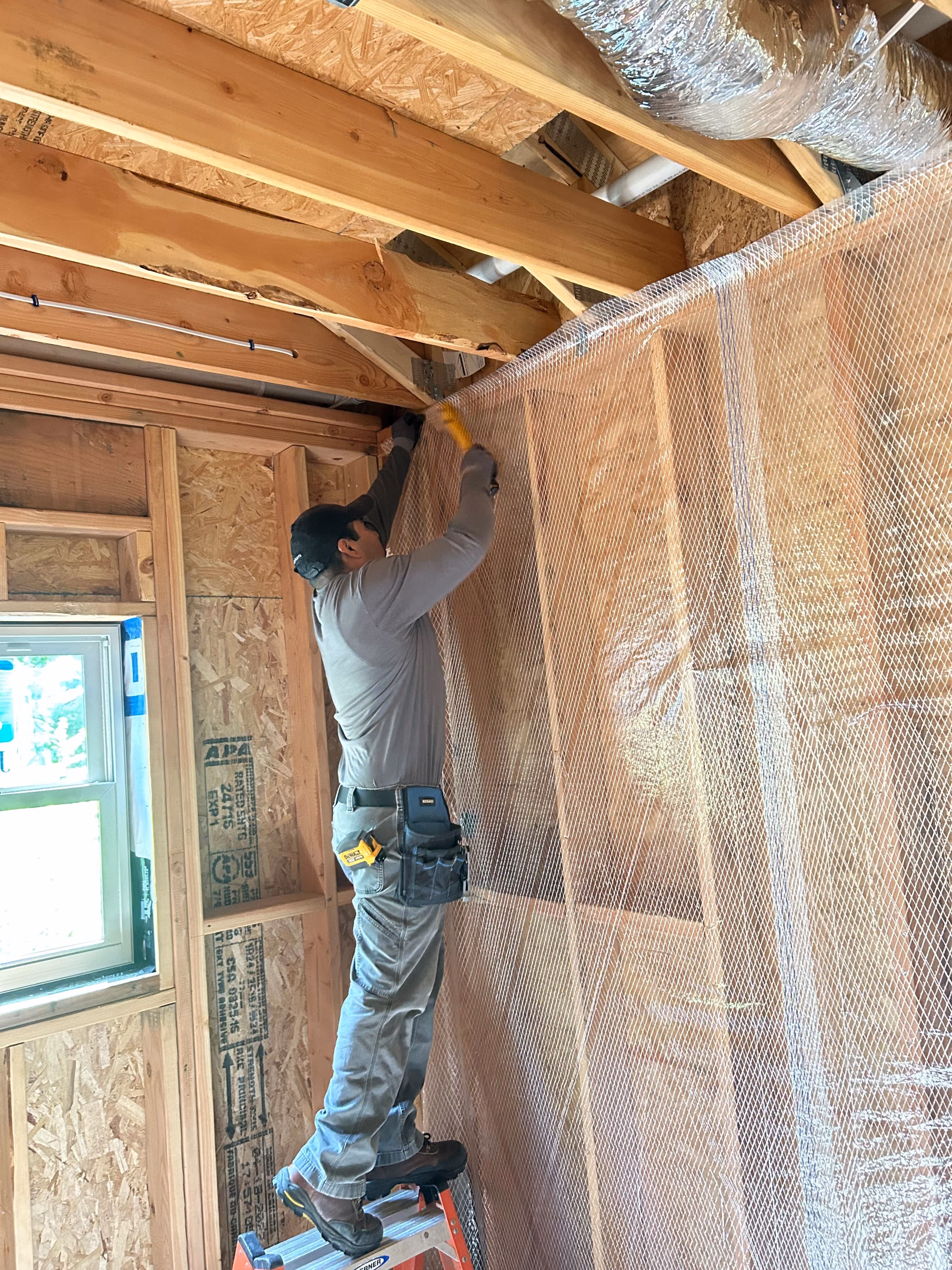 Construction worker wearing gloves and a cap installing mesh netting on wooden framing inside a building under construction.