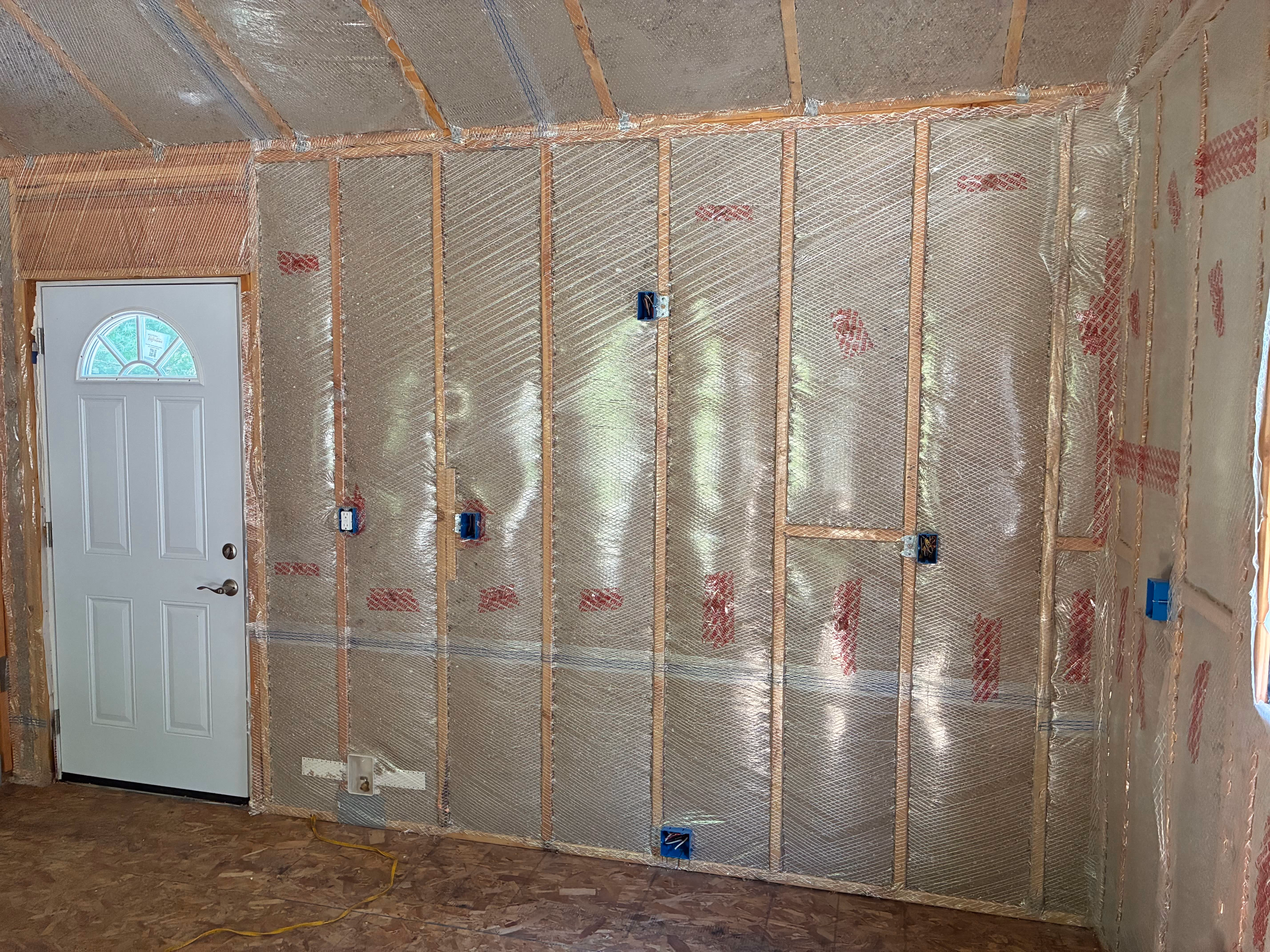 Interior wall under construction with wooden studs and reflective insulation installed, next to a white door with a semi-circular window.