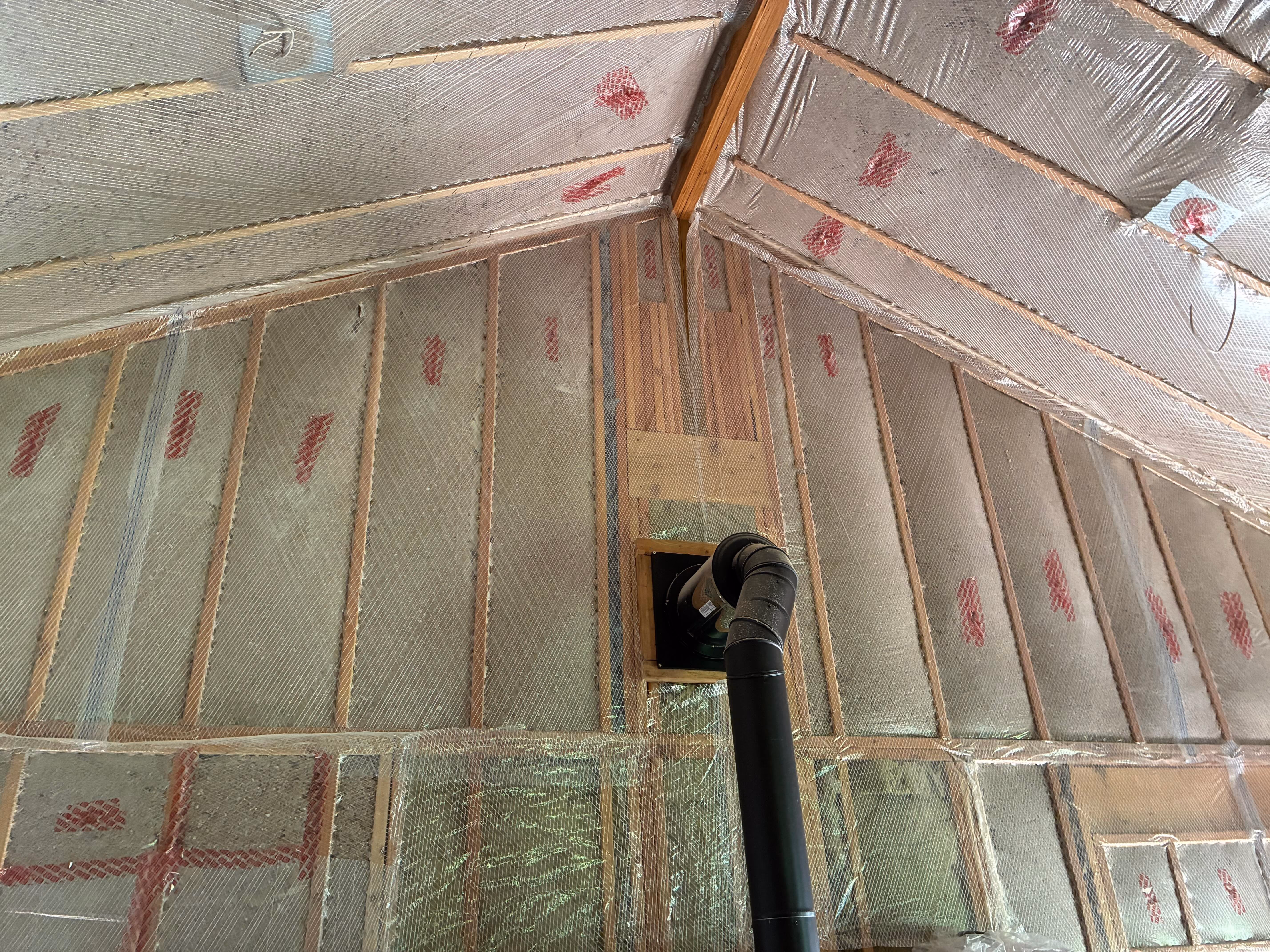 Interior wall and ceiling framed with wooden studs, covered with reflective foil insulation and a black vent pipe running upwards.