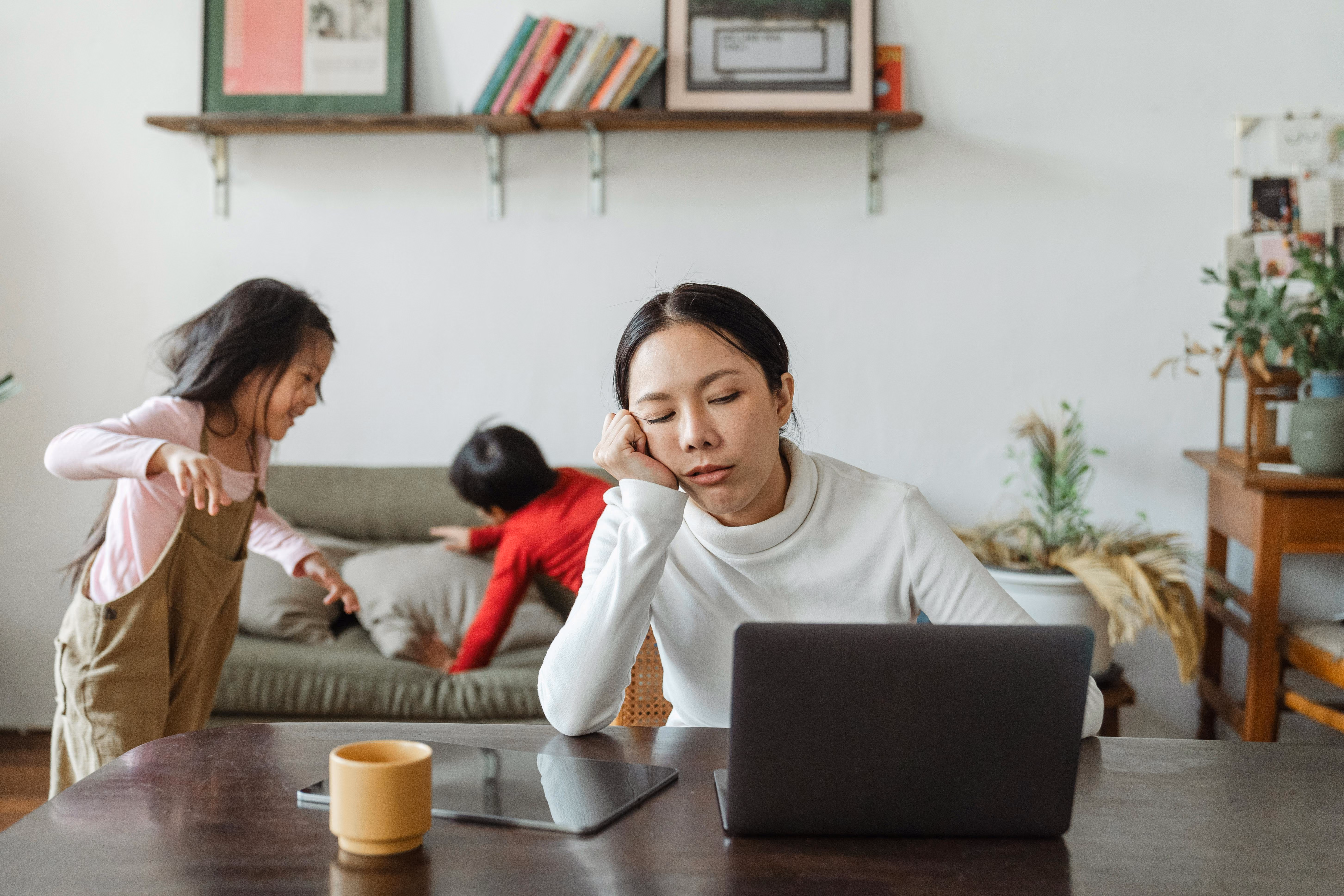 Tired woman resting her head on her hand at a laptop while two children play energetically on a couch behind her.