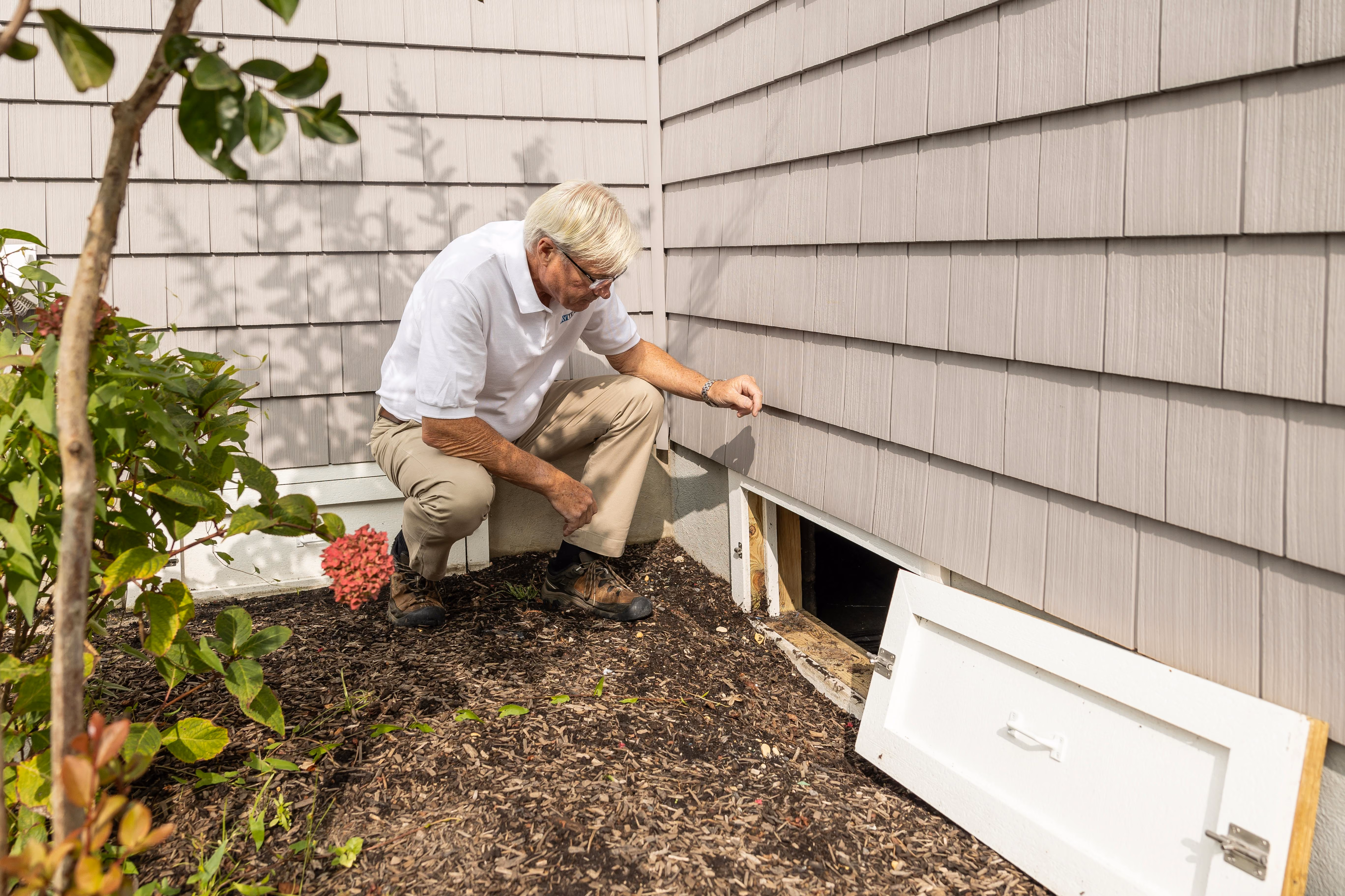 Man in a white shirt crouching outside a house, inspecting an open crawl space door near the foundation.