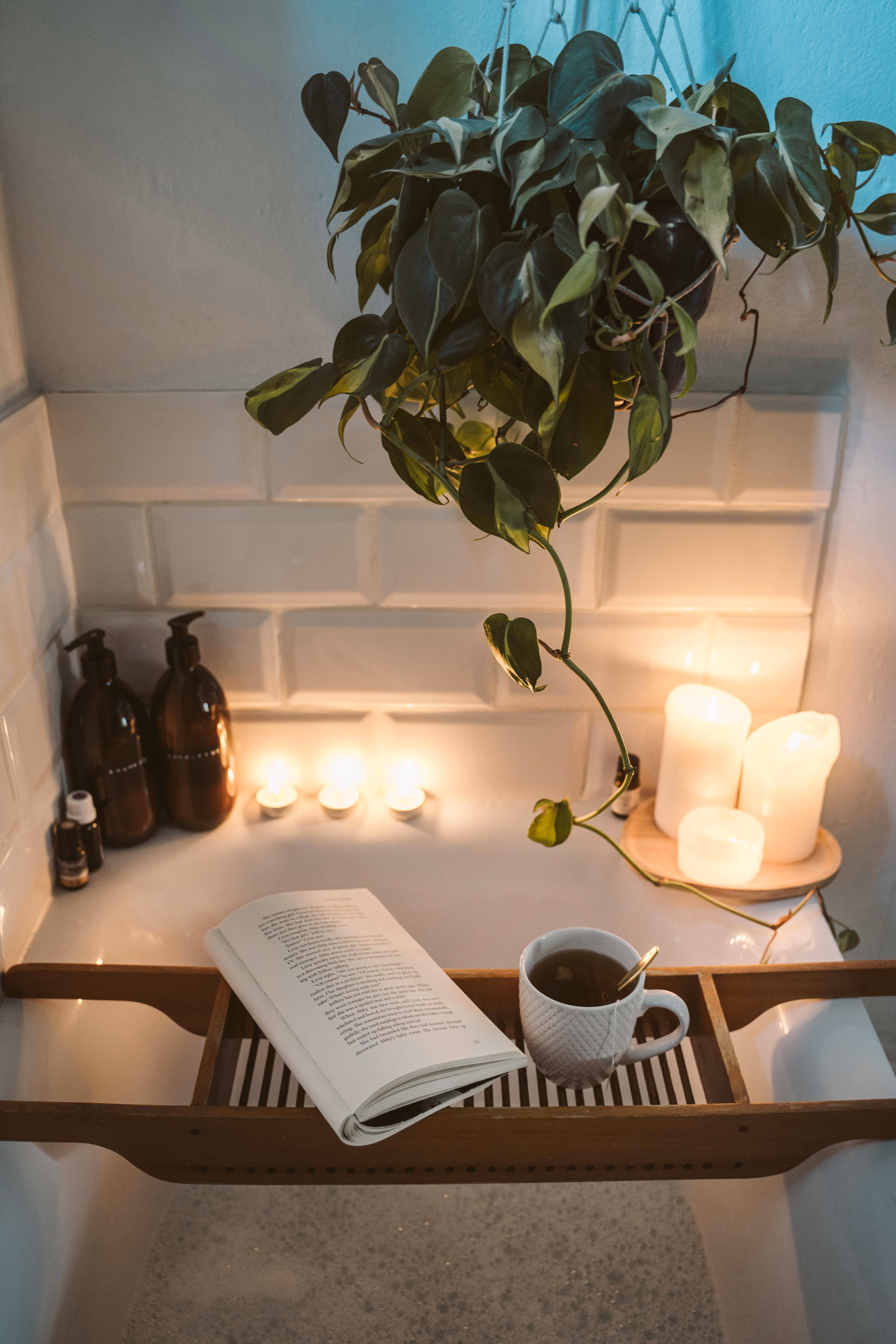 Relaxing bathtub scene with a wooden tray holding an open book and a cup of tea, surrounded by lit candles and a hanging green plant.