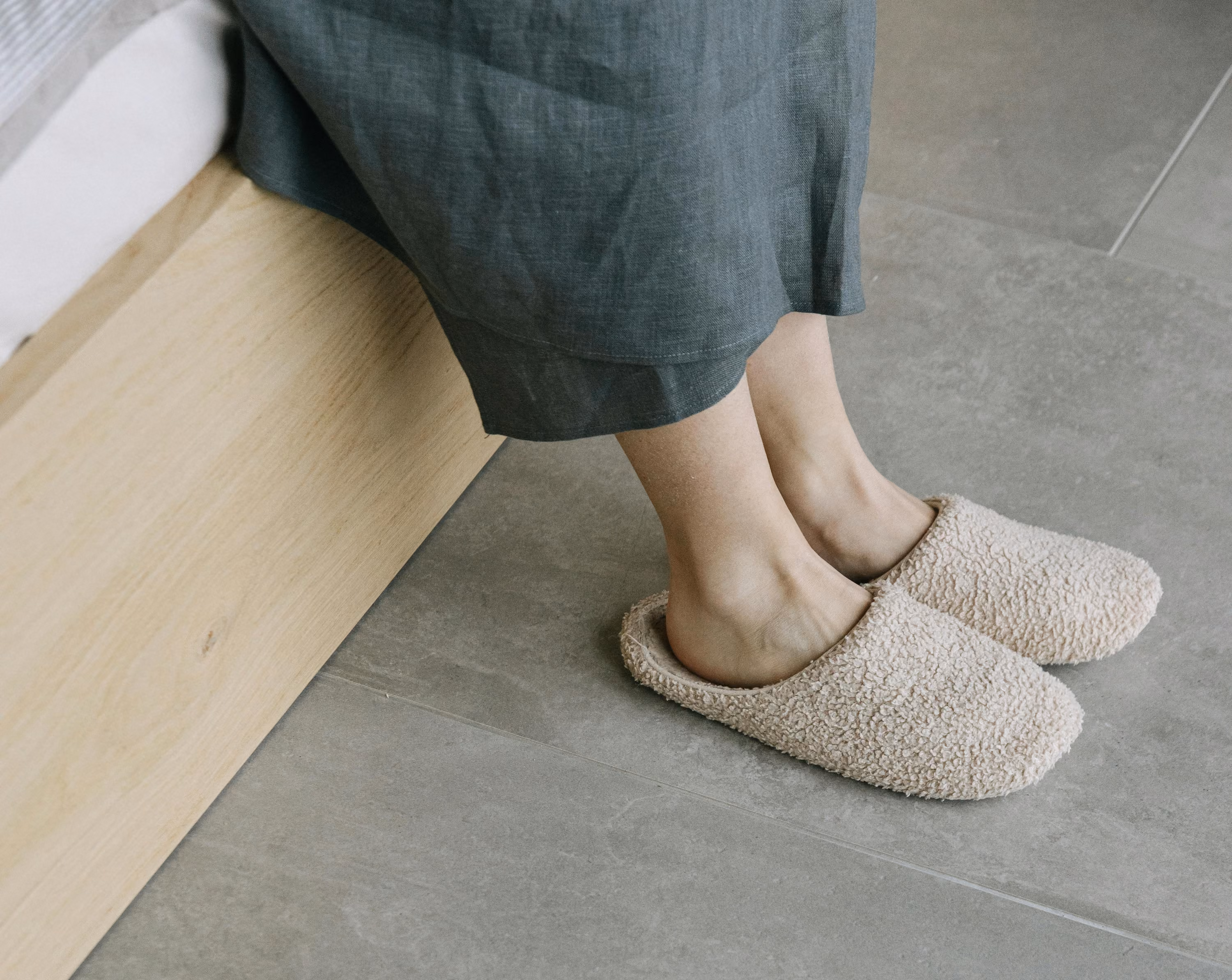 Person sitting on a wooden bed frame with feet wearing beige fuzzy slippers on gray tiled floor.