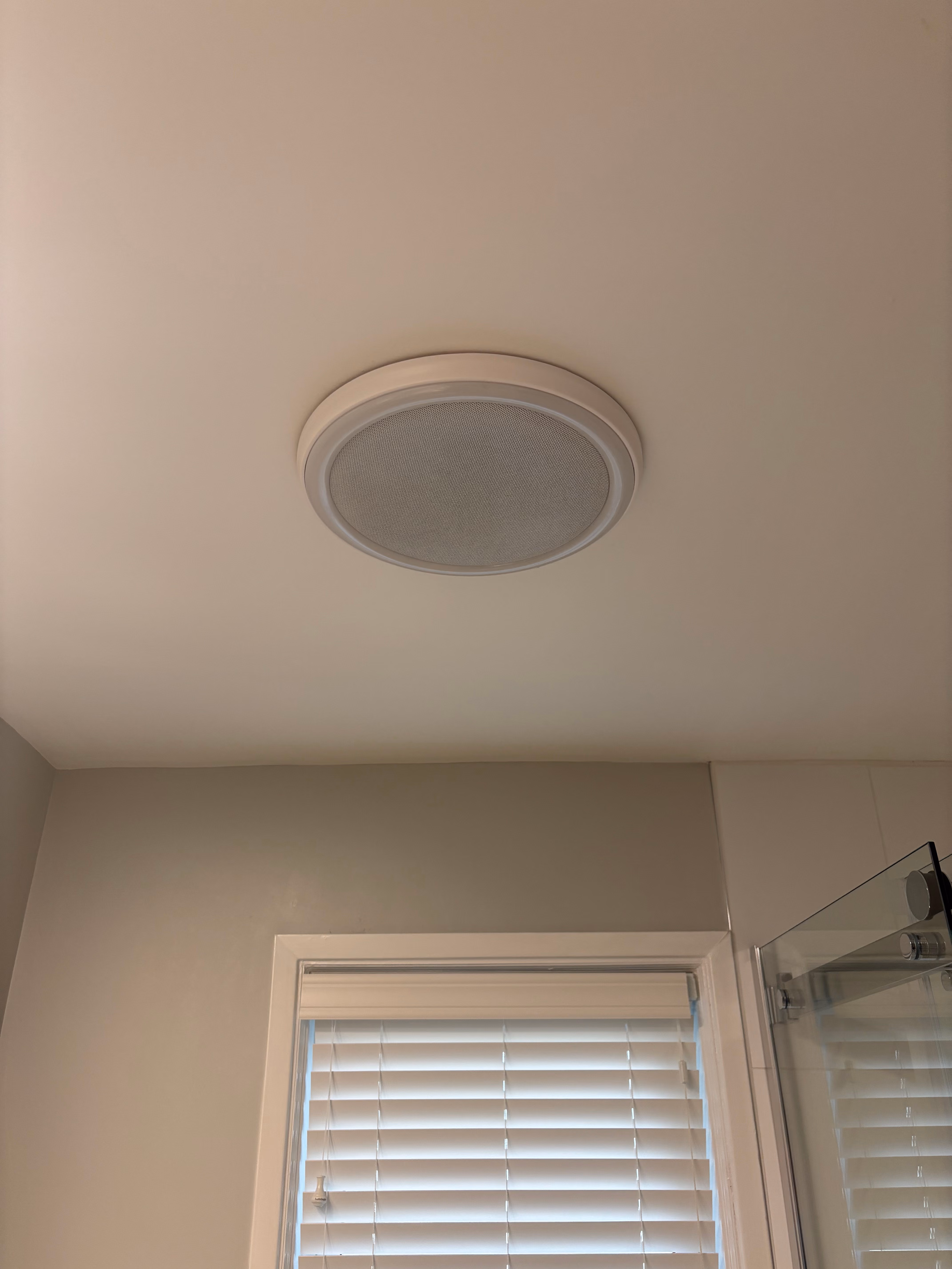 White circular ceiling exhaust fan installed on a beige bathroom ceiling above a window with closed blinds and a glass shower door.