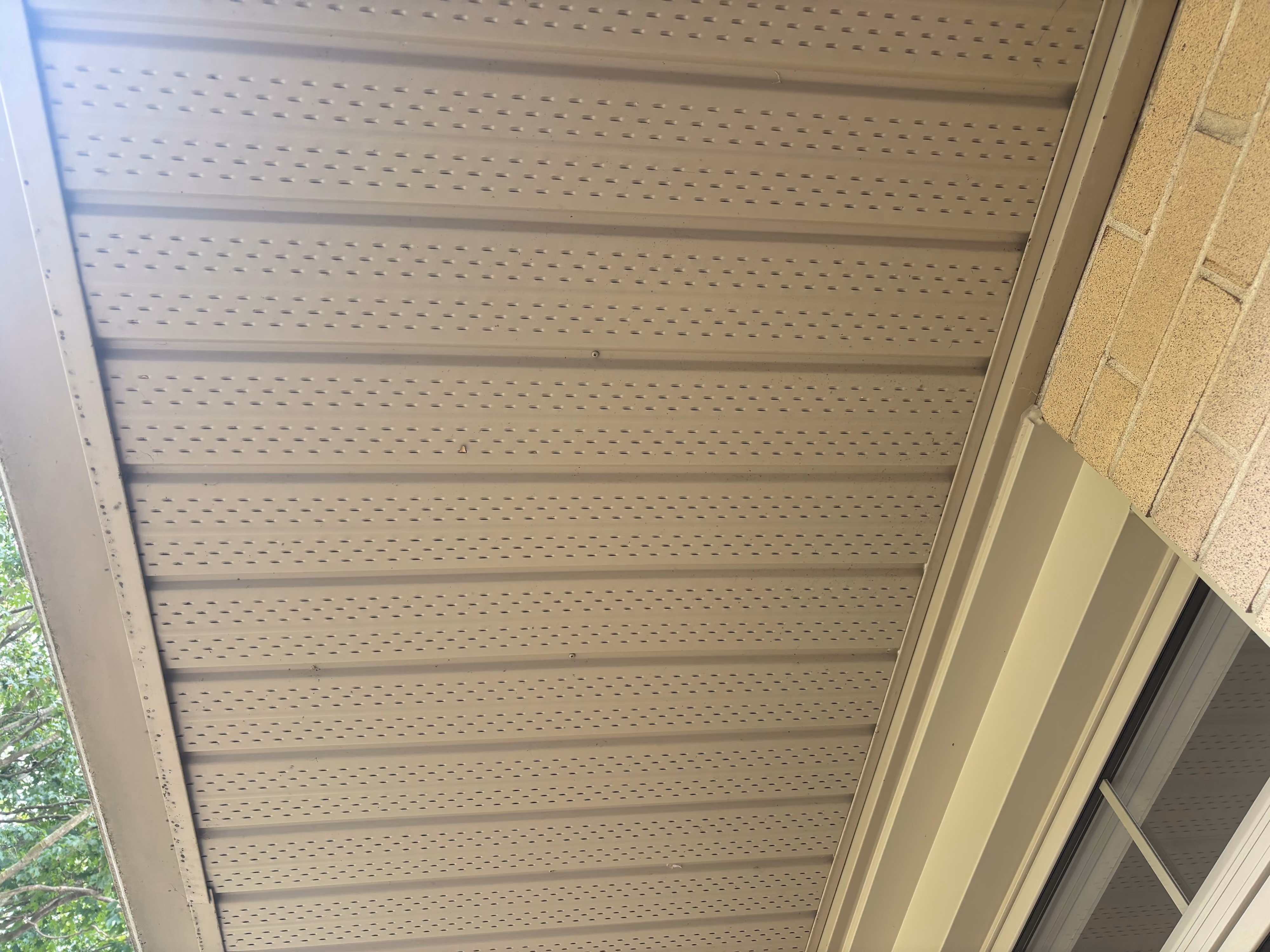 Beige perforated soffit panels installed beneath the roof edge of a building with a brick wall and window.