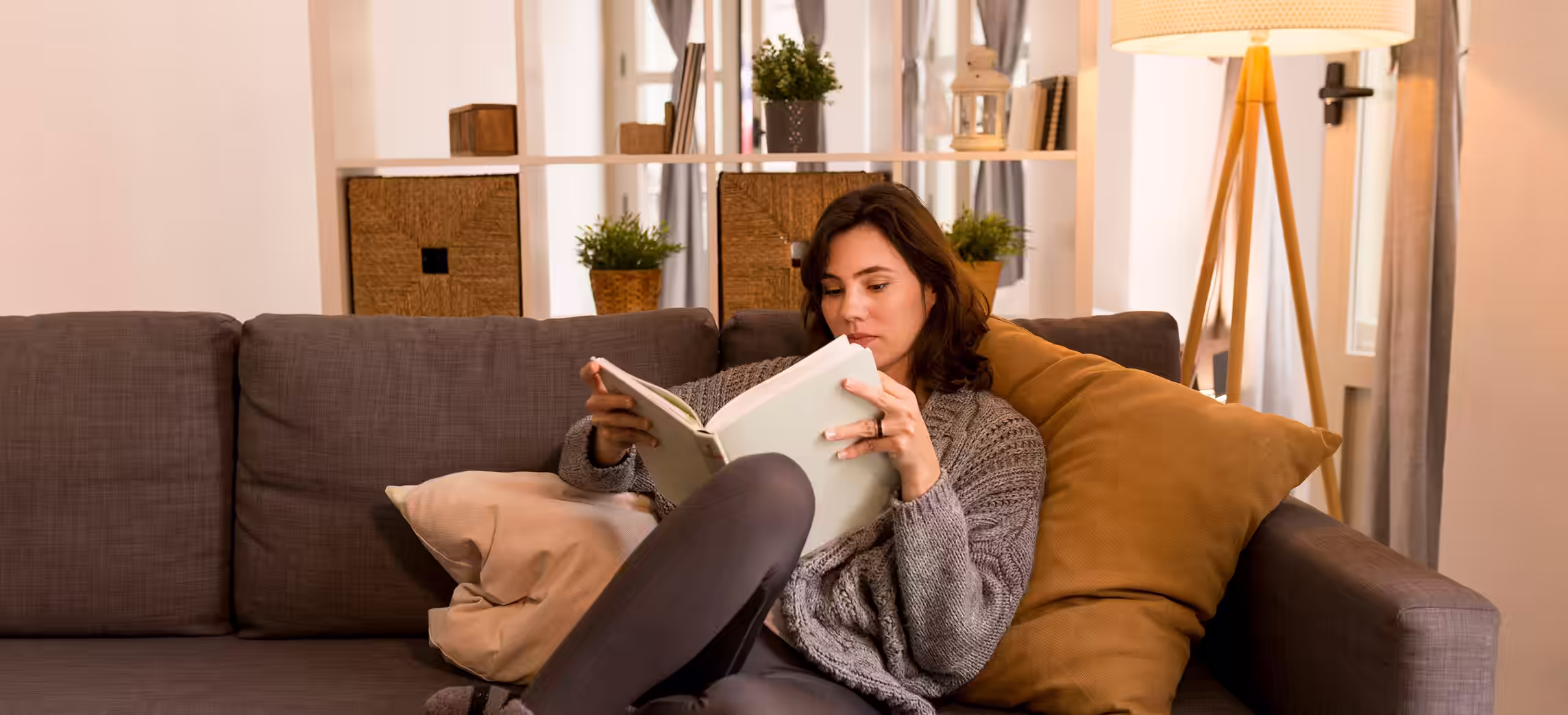 Woman in gray sweater reading a book while sitting on a gray couch with beige and mustard pillows in a cozy living room.