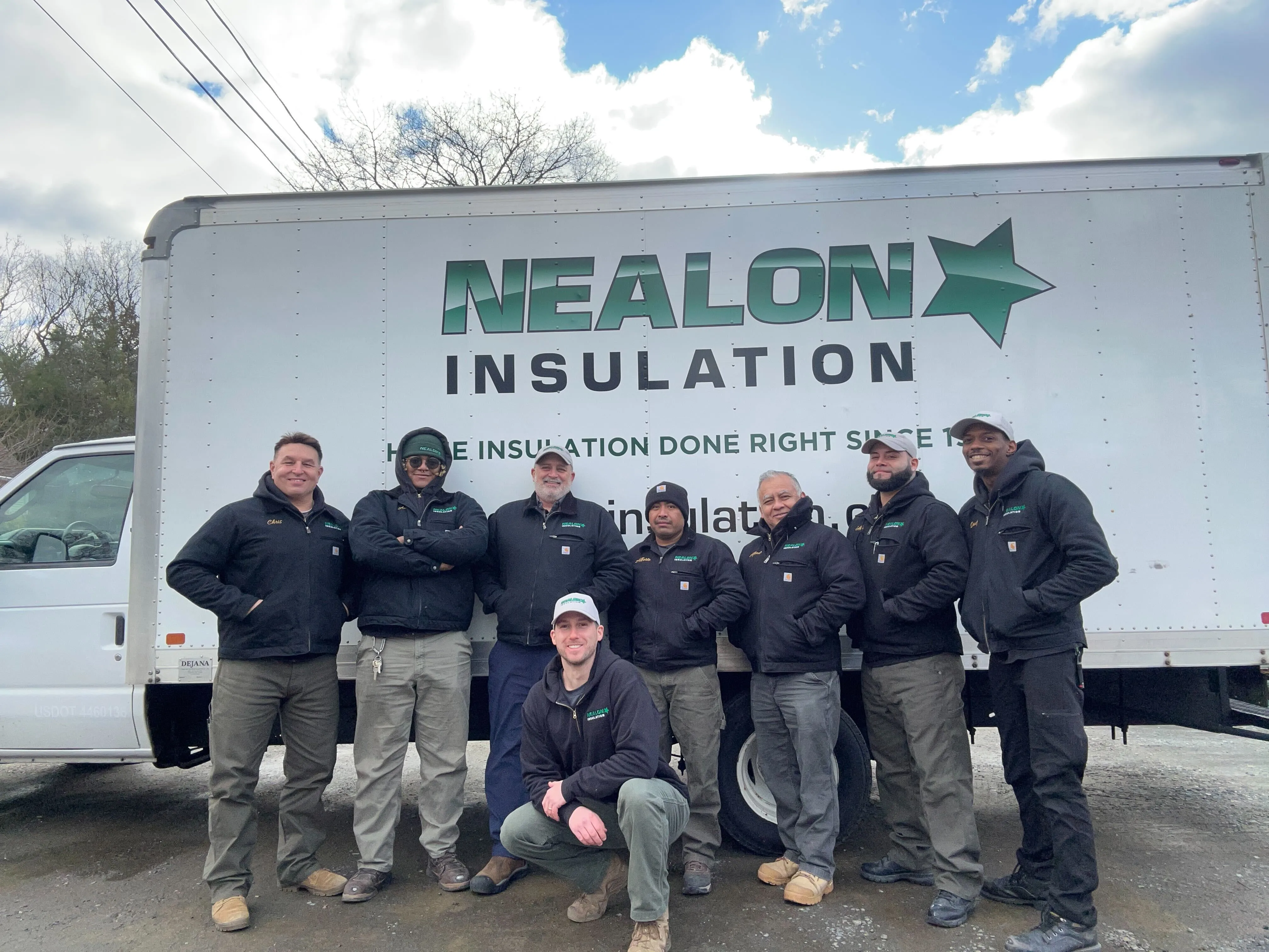 Group of eight workers wearing black jackets posing in front of a Nealon Insulation company truck.
