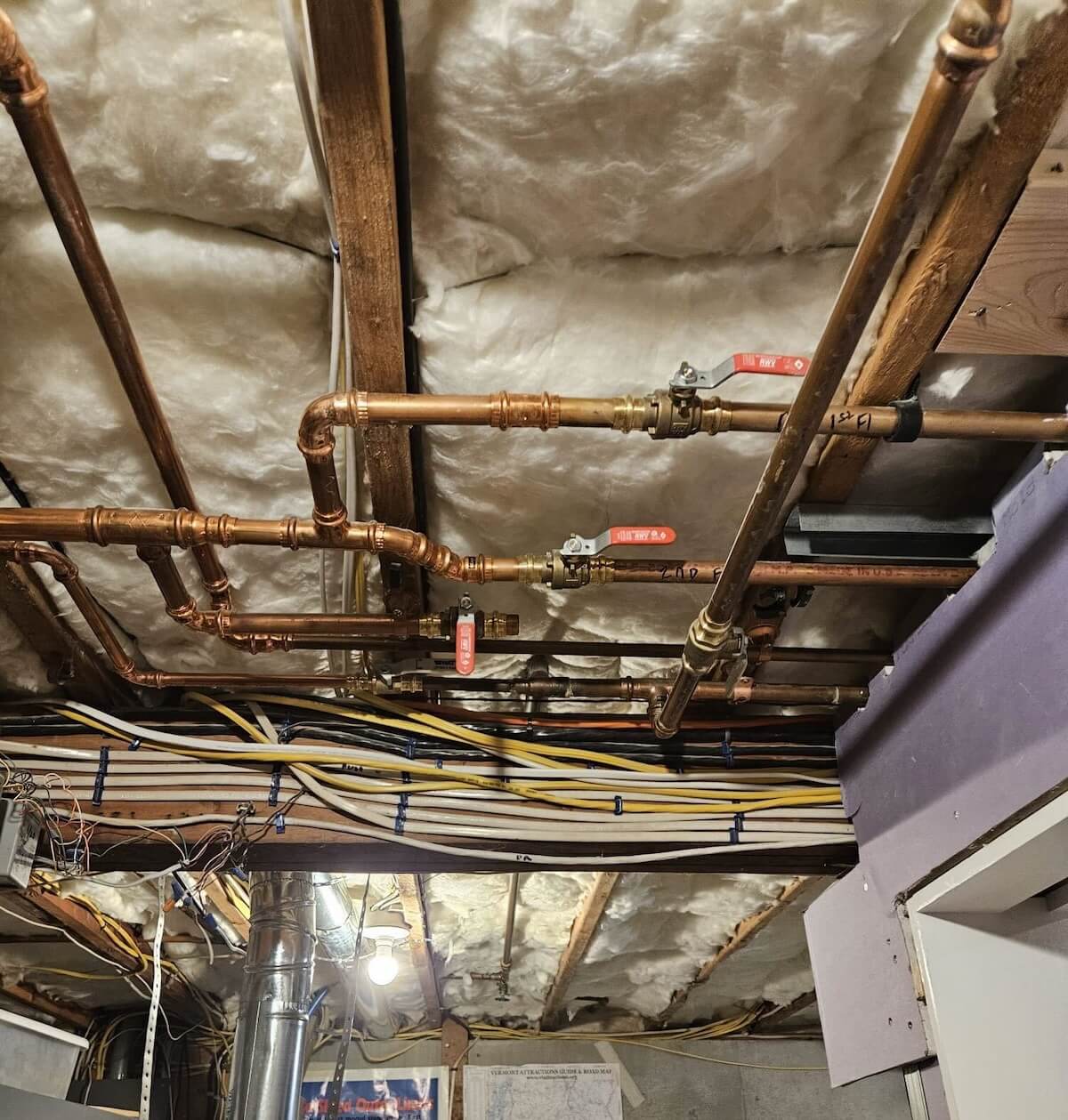 Basement ceiling with fiberglass insulation, copper pipes with valves, and organized electrical wiring.