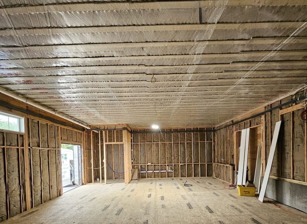 Interior of a room under construction with exposed wooden framing and wall insulation, ceiling covered with reflective insulation, and construction materials on the floor.