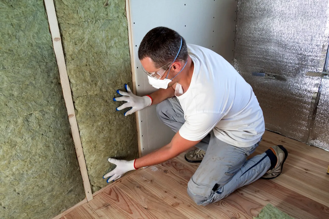 Worker wearing protective goggles, respirator mask, and gloves, installing rock wool insulation inside a wooden frame wall.