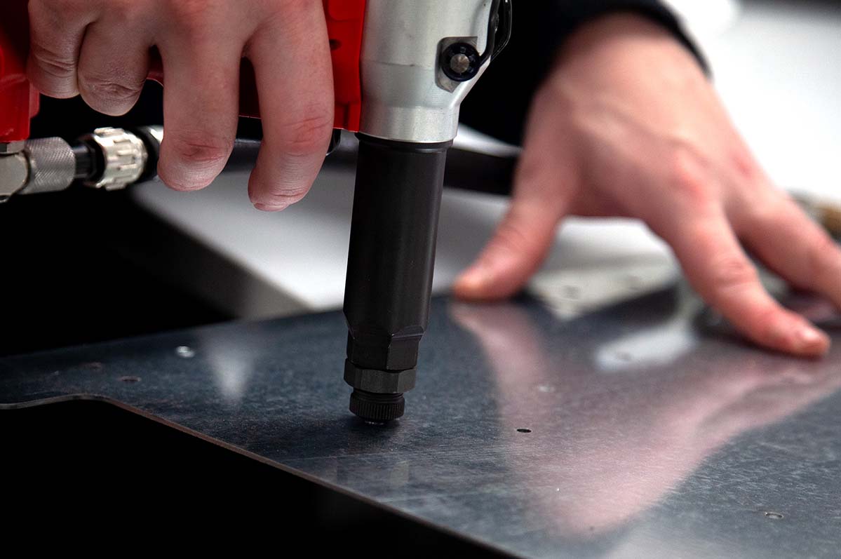 Technician using a pneumatic rivet tool to secure a metal plate, showcasing hands-on fabrication work during the panel or wiring enclosure build process at Tec-Stop’s UK facility.