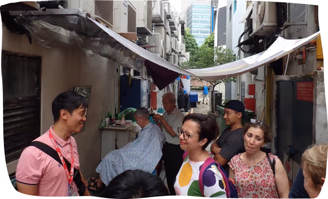 People gathered in a narrow alley where a man is giving a haircut to an elderly person seated on a chair.