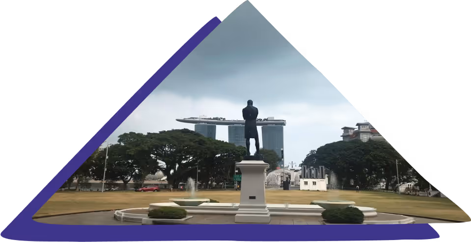 Statue of a man on a pedestal in front of gardens and fountains with Marina Bay Sands hotel in the background under a cloudy sky.