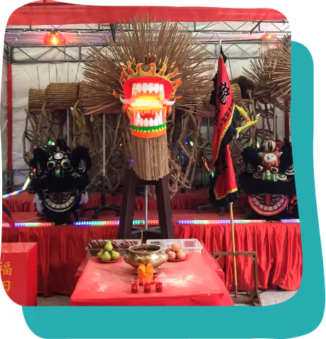 Traditional Chinese dragon head made of straw and wood with a red and white face, displayed on a stand with offerings of fruit and candles on a red table in front.