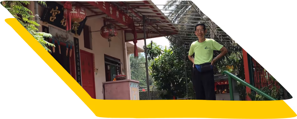Man in a green shirt standing and smiling near a traditional building with red lanterns and greenery.