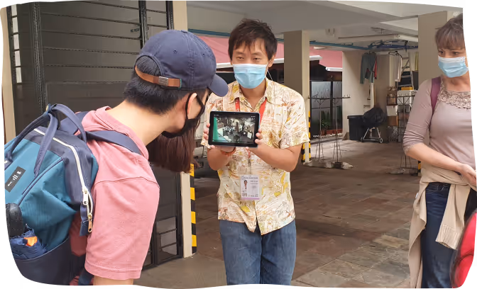 Man wearing a face mask showing a tablet screen to a small group of people outdoors near a building entrance.