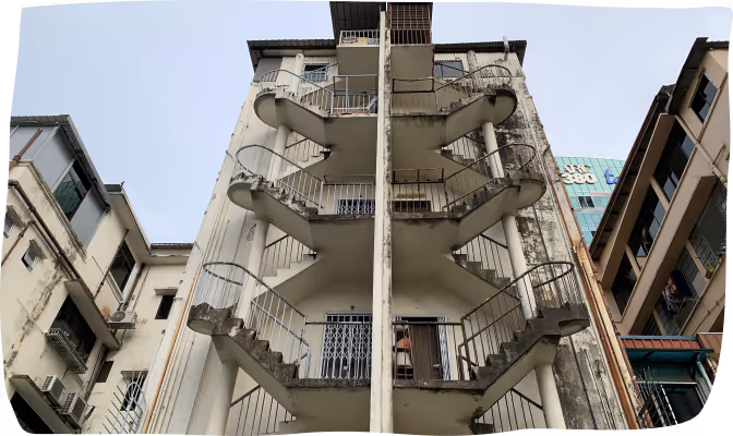Upward view of an old concrete building with external staircases on both sides and adjacent buildings in an urban setting.