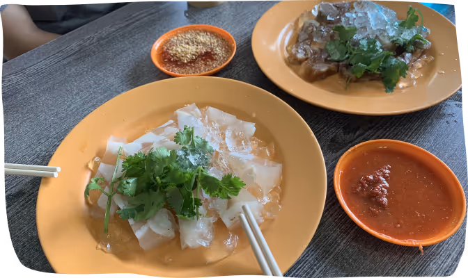 Two orange plates on a table with chilled rice noodle rolls garnished with cilantro and ice, accompanied by small bowls of chili sauce and crushed peanuts.