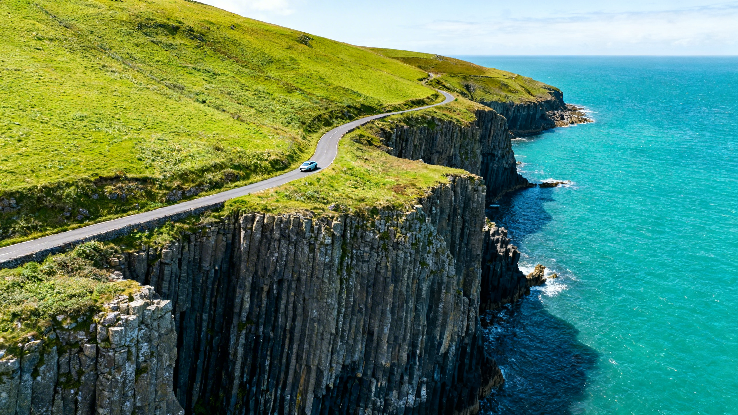 The Antrim Coast Road in Northern Ireland, basalt sea cliffs dropping to a vivid turquoise sea, a narrow coastal road winding along the cliff edge