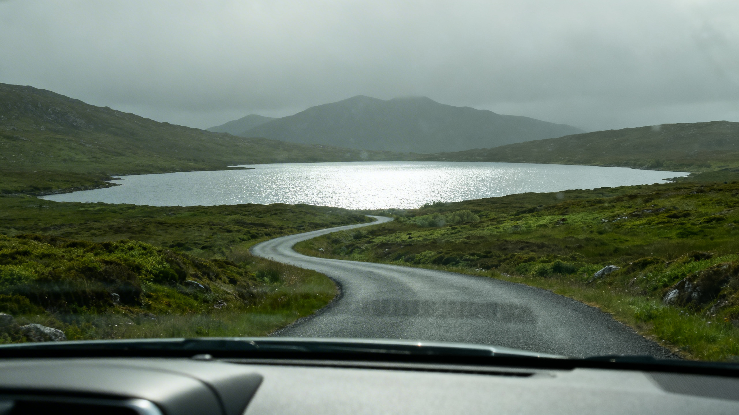 View through a car windscreen of a winding road through the Connemara bog landscape, grey-green hills in the distance under a soft overcast sky