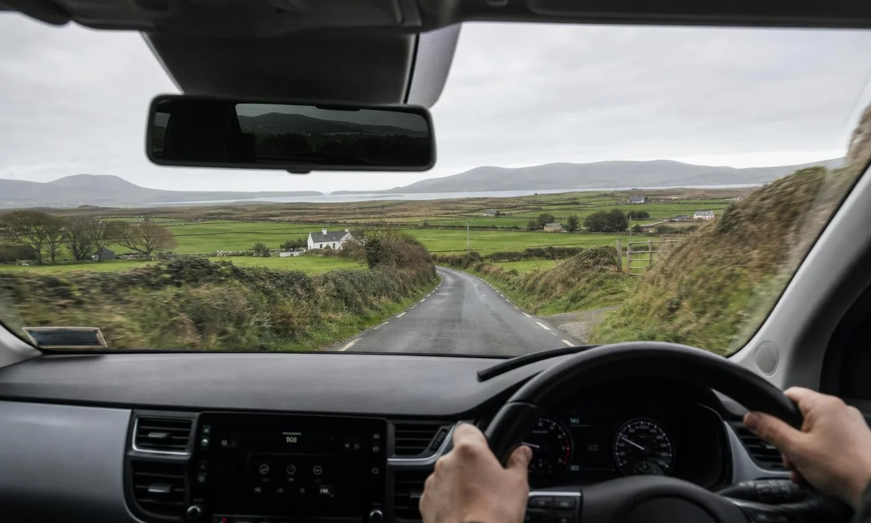 Driver's view from behind the wheel of a modern car on an Irish country road, left-hand driving position visible, green fields ahead. No branding. Natural light. Documentary DSLR style.