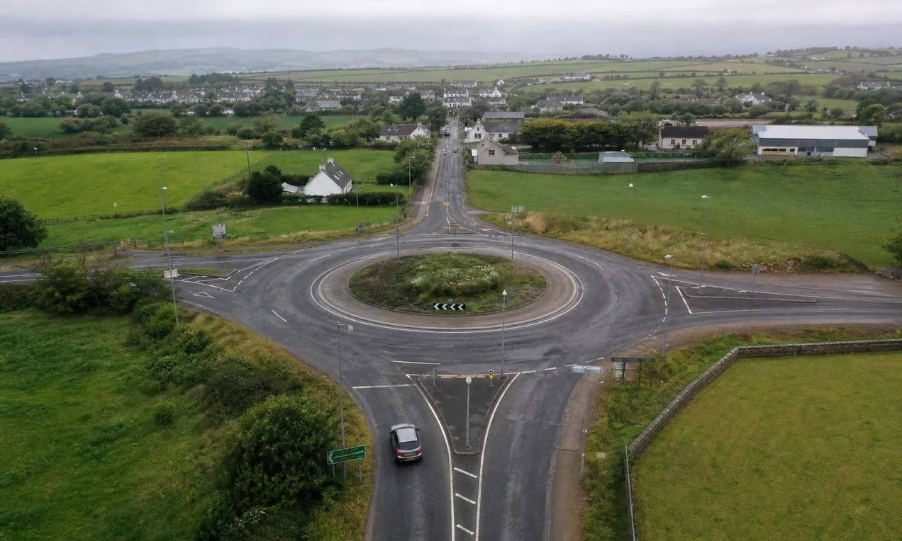 Aerial view of a rural Irish roundabout with a car entering, green fields and a small village in the background. Overcast day. No visible car branding.