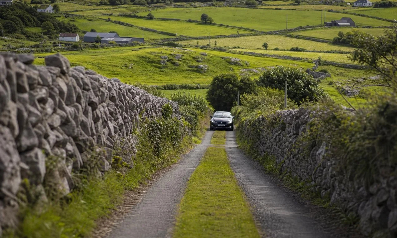 Narrow Irish boreen with grass growing down the centre, stone walls on both sides, a car approaching in the distance. Green landscape. No branding. Natural light DSLR.