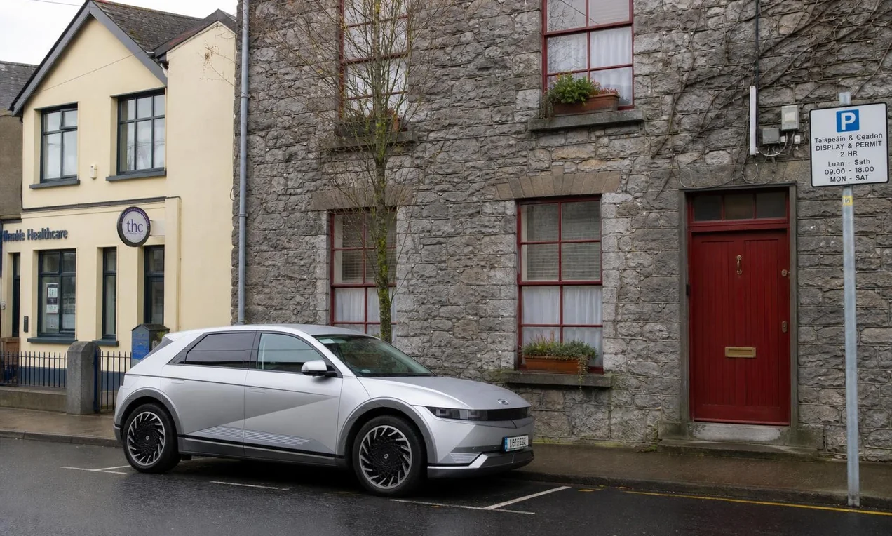 Modern car parallel parked on a narrow street in an Irish town, stone buildings behind, parking sign visible on a pole. Overcast day, no decals. Documentary style.
