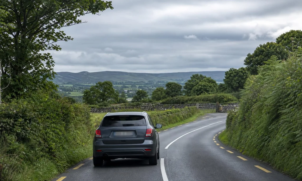 Modern car driving on a winding Irish R-road with green hedgerows on both sides, stone walls in the distance, overcast sky.