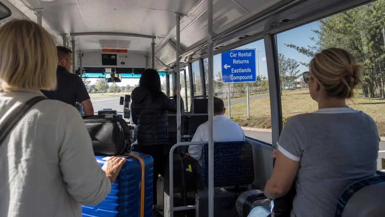 Interior of an airport shuttle bus with passengers holding their luggage, a sign for Car Rental Returns visible through the window.