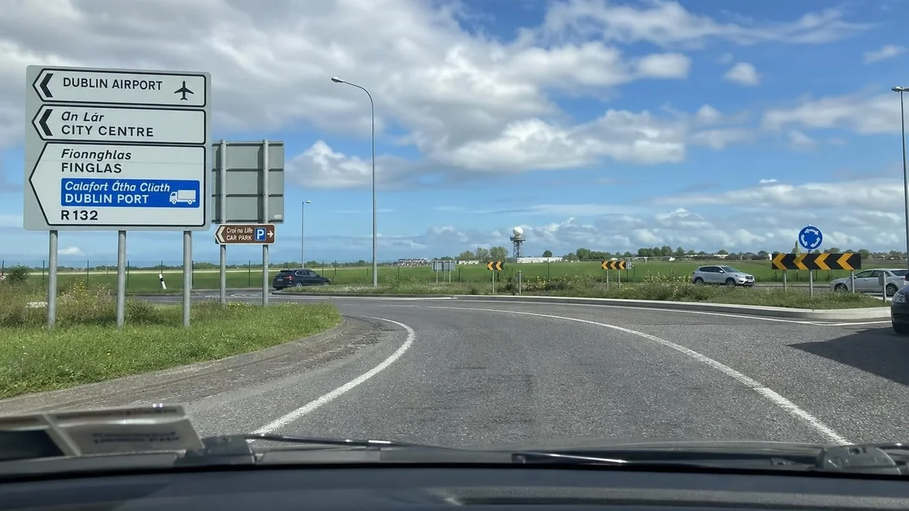 A view through a car windscreen approaching a roundabout near Dublin Airport with road signs clearly visible and green countryside in the distance.