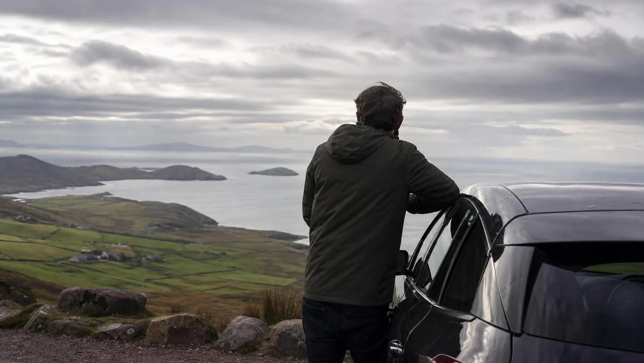 A relaxed driver looking out over a scenic coastal view in Kerry, the car parked at a viewpoint, Atlantic Ocean in the distance. Back view, no faces. Dramatic overcast sky.