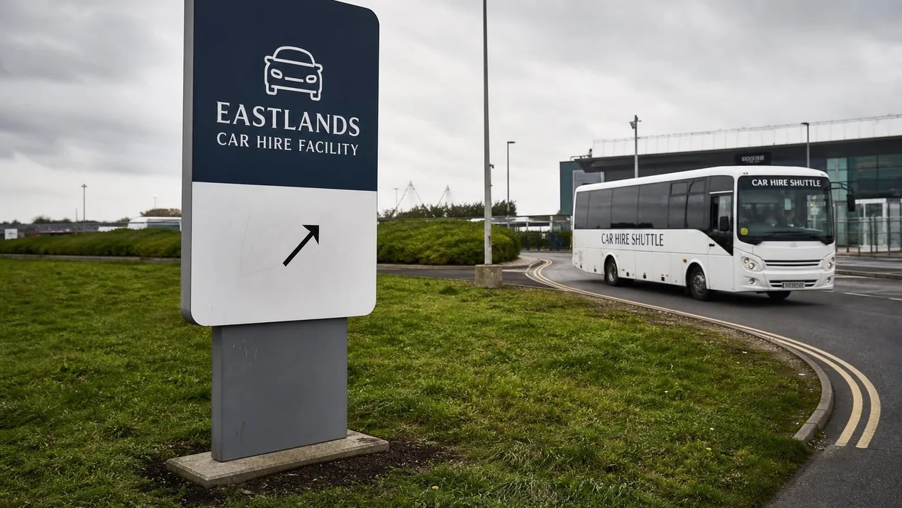A sign for the Eastlands Car Hire Facility near Dublin Airport with a shuttle bus visible in the background. Overcast day. Documentary style.