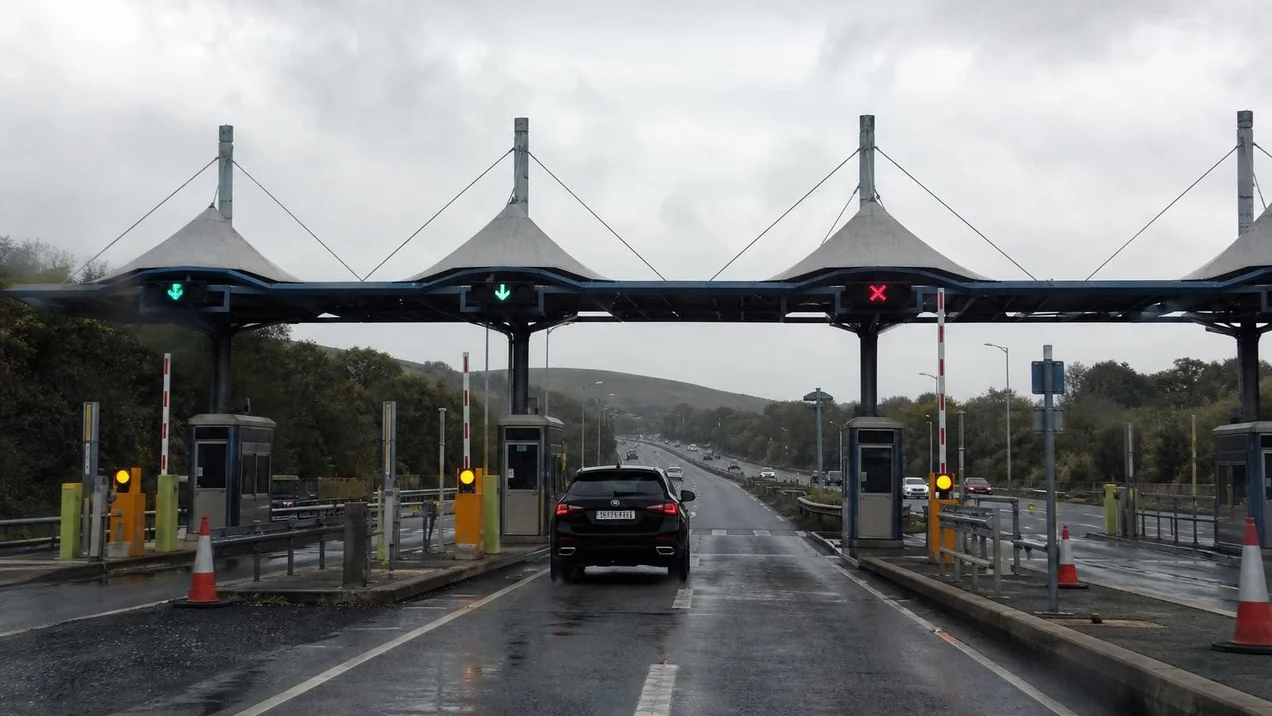 An Irish toll plaza on a motorway, a car approaching the barrier, overcast sky. No identifiable branding. Documentary style.