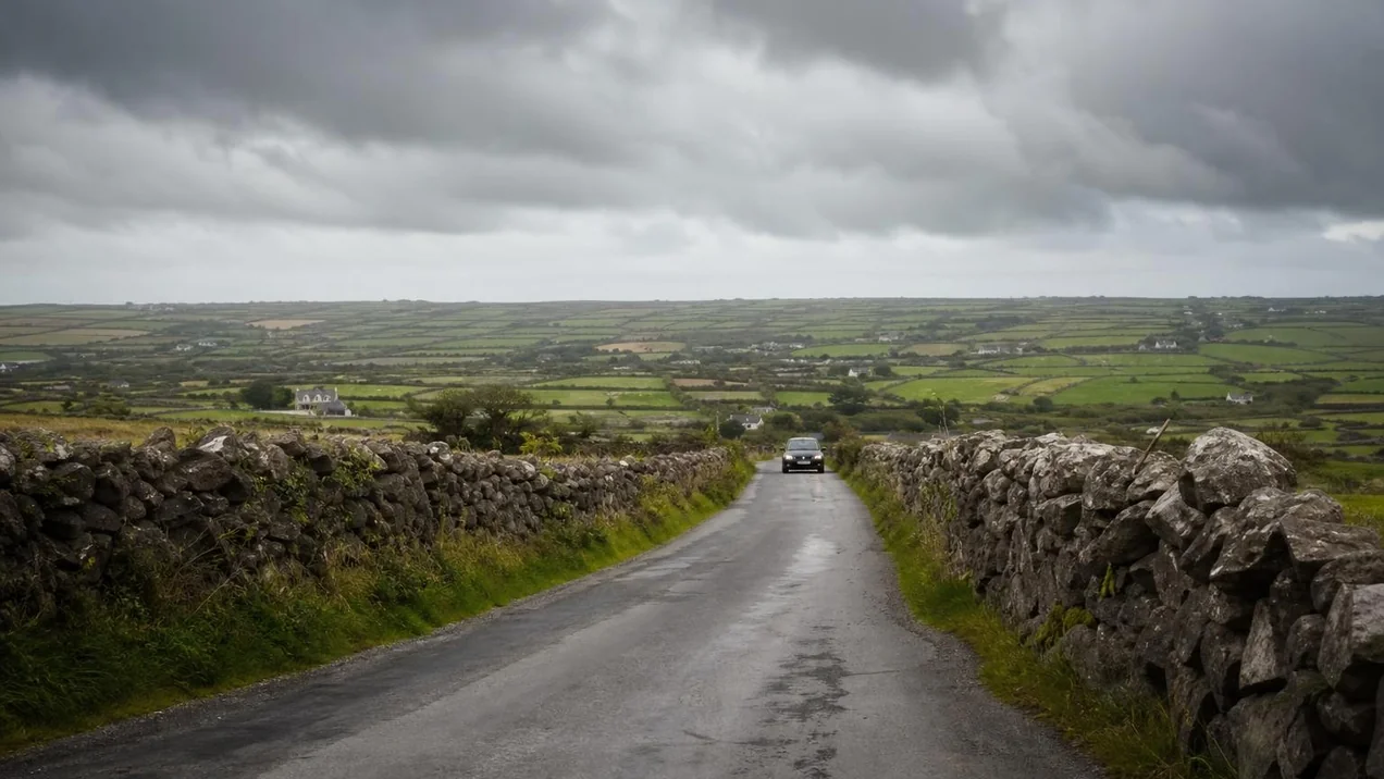 A narrow Irish country road with stone walls on both sides and a car approaching in the distance, green fields beyond. Overcast day. Documentary style.