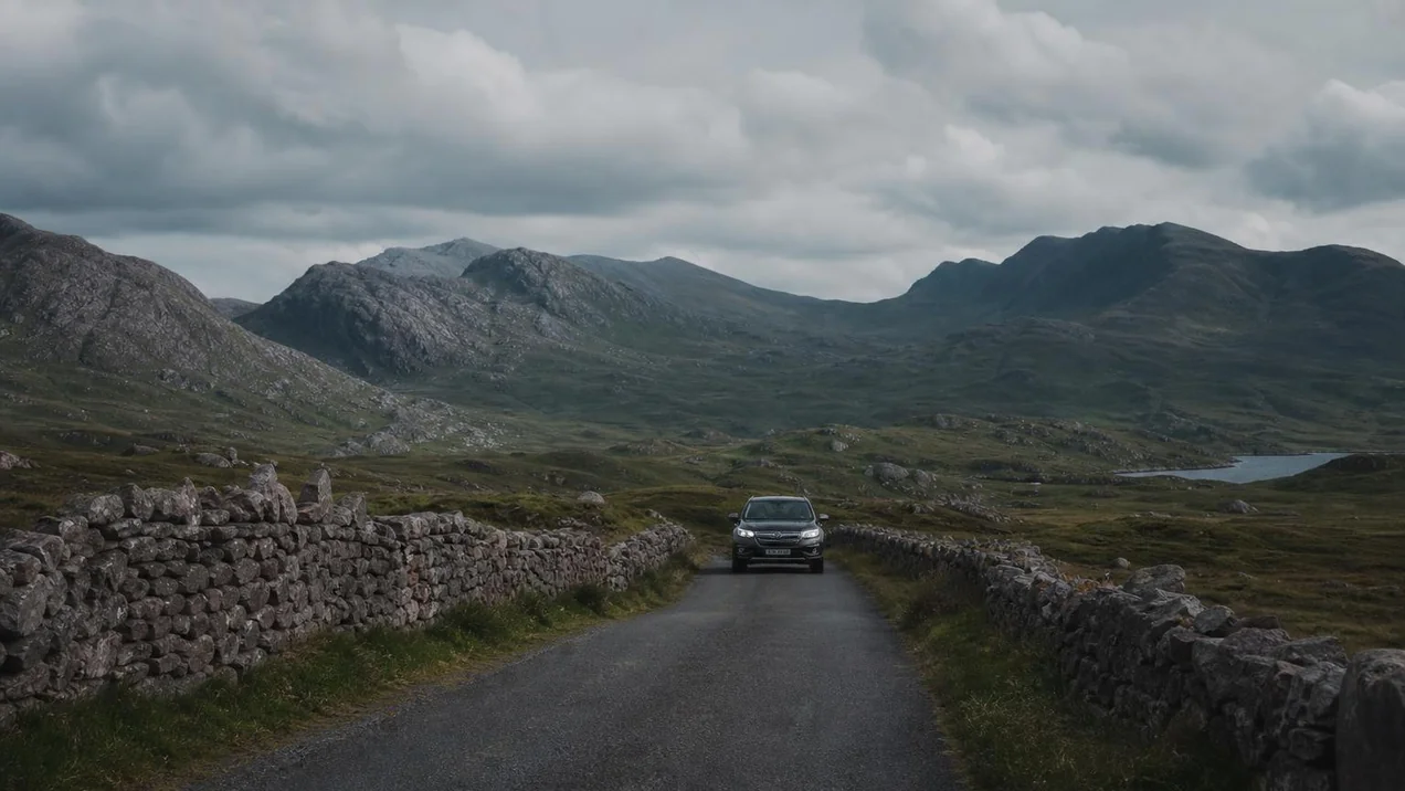 A narrow country road in Connemara with stone walls on both sides, green hills in the distance, a car approaching. Overcast dramatic sky. Documentary style.