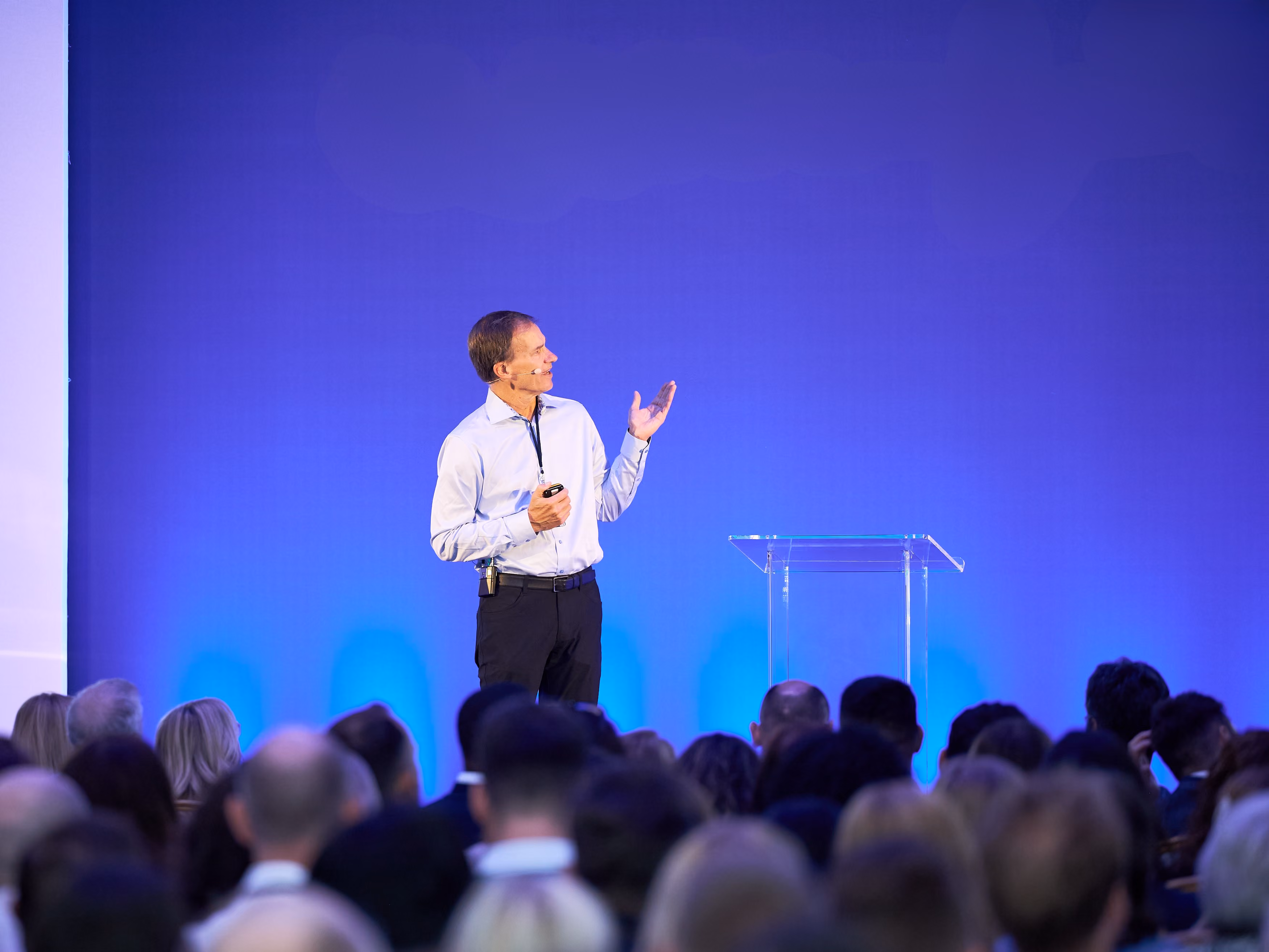 A speaker presenting to a large seated audience on a blue-lit conference stage